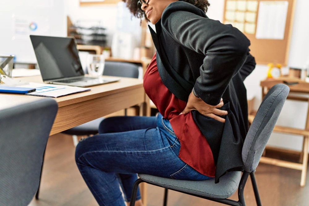 dolore-lombare-destro-Donna in ufficio seduta alla scrivania con laptop, mano sulla schiena per un dolore lombare durante il lavoro